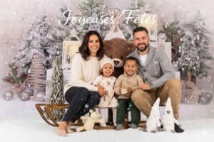 Famille souriante de quatre personnes (parents, une petite fille et un petit garçon) posant sur un traîneau en bois dans un décor de Noël enneigé avec sapins givrés et décorations lumineuses. Le texte "Joyeuses Fêtes" est inscrit en blanc au centre supérieur de l'image. Photo de séance de Noël réalisée par Alex, photographe professionnel à Vesoul.